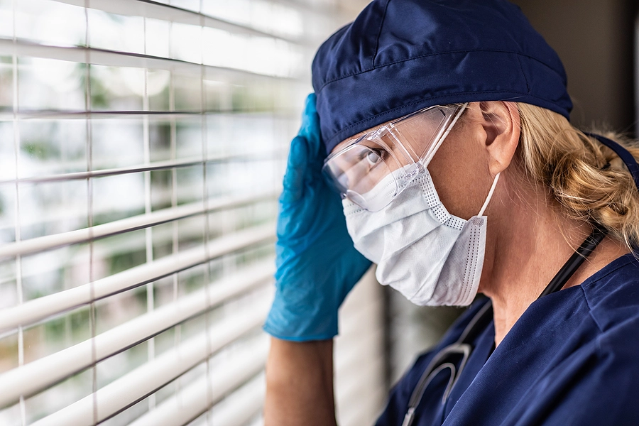 Female health professional wearing facemask and goggles looking through blinds with worried expression, hand on forehead