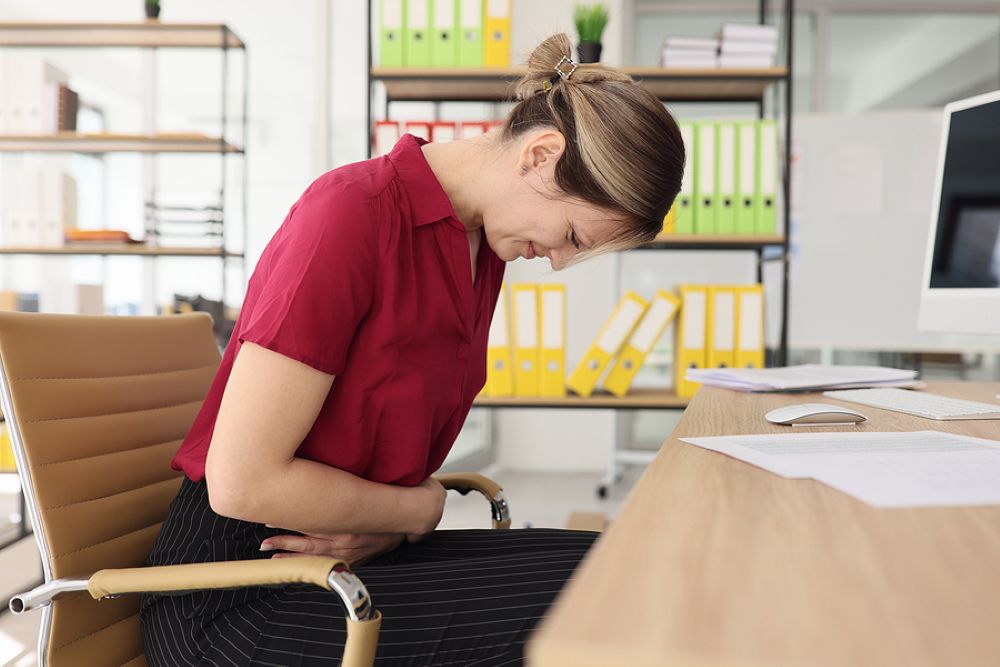 Woman in office with severe stomach ache clutching belly while seated at desk