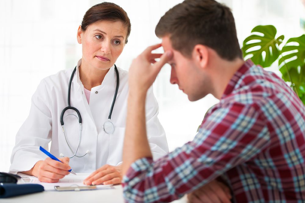 Woman doctor wearing a stethoscope is talking with a man wearing a plaid shirt who is resting his head on his hand worried.