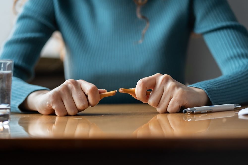 Woman wearing a blue sweater, close up of her hands on a desk snapping a pencil in two