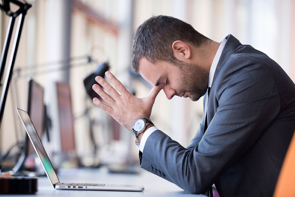 Businessman in suit at desk with hands on head having a migraine headache