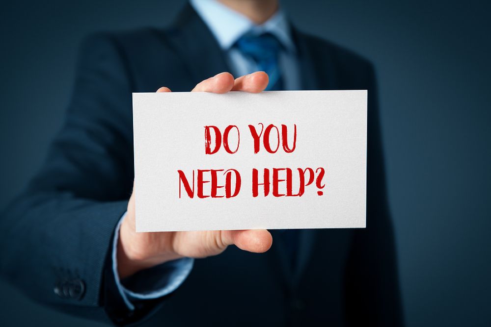 A man in a business suit is holding a card that reads "Do You Need Help" printed in red capital letters